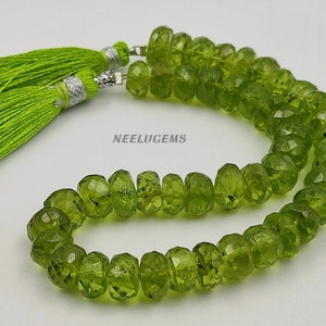 Op de afbeelding: Een ketting van gefacetteerde, olijfgroene peridot kralen met aan elk uiteinde een heldergroene kwast. De kwasten zijn gemaakt van fijne draden en hebben zilverkleurige doppen. De kralen zijn gerangschikt in een ketting- of armbandstijl.