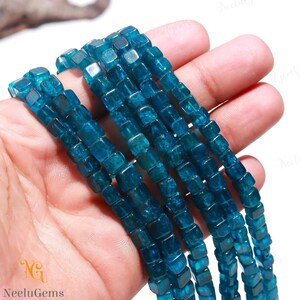 Puede incluir: Una hebra de cuentas de apatita azul turquesa, cada cuenta tiene forma cuadrada. Las cuentas están enhebradas en un cordón elástico transparente y se muestran en una mano.