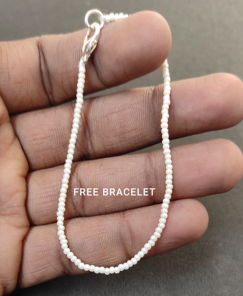 May include: A white beaded bracelet with a silver clasp. The bracelet is on a person's hand. "FREE BRACELET" is written on the hand.