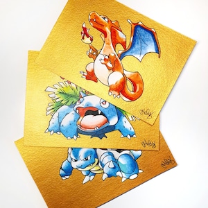 Può includere: Tre illustrazioni ad acquerello di personaggi Pokémon: Charizard, Venusaur e Blastoise. Ogni personaggio è raffigurato su un quadrato dorato strutturato. Charizard è arancione e rosso con ali blu. Venusaur è blu e verde. Blastoise è blu.