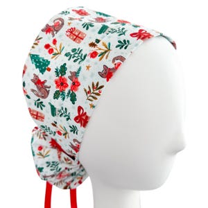 May include: A white scrub cap featuring a Christmas-themed pattern. The design includes red foxes, gift boxes, Christmas trees, and floral accents. The cap has red ties and is displayed on a mannequin head.