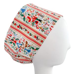 May include: A white and red patterned scrub cap featuring a Christmas-themed design. The cap has a repeating pattern of nutcrackers, angels, and other holiday elements. Red and gold stripes run across the cap.