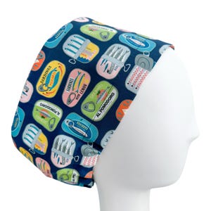 May include: A navy blue surgical scrub cap featuring a repeating pattern of colorful sardine cans. The cans are in various colors and designs, with text labels like "Sardines with Lemon" and "Al Pomodoro". The cap is designed to fit comfortably.