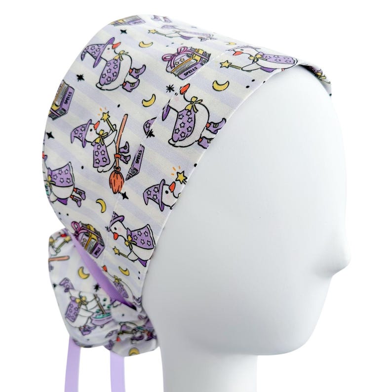 May include: A white scrub cap with a repeating pattern of cartoon witch characters, stars, moons, and spell books. The cap has a purple striped background and purple ties.