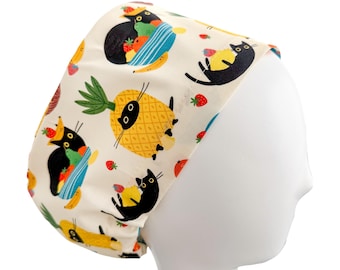May include: A cream-colored surgical scrub cap featuring a repeating pattern of black cats, strawberries, bananas, and pineapples. The cat illustrations have yellow eyes and are surrounded by colorful fruit. The cap is designed to fit the head and is displayed on a mannequin head.