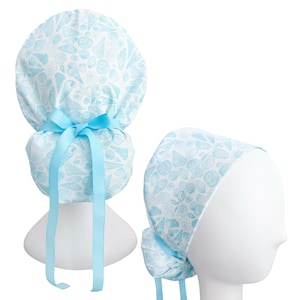 May include: A light blue and white patterned scrub cap featuring a seashell design. The cap is tied with a light blue ribbon. The cap is displayed on a white mannequin head.