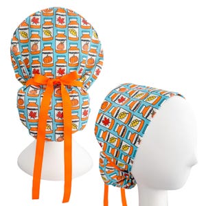 May include: A blue surgical scrub cap with an orange ribbon. The cap is patterned with orange jars, pumpkins, and fall leaves. The text "PROPOSEALL" is printed on the jars. The cap is displayed on a white mannequin head.