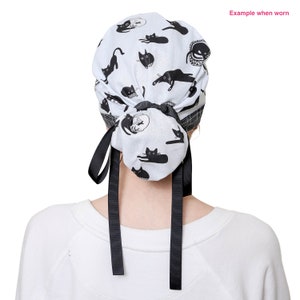 May include: A white scrub cap featuring a repeating pattern of black cat illustrations. The cap has a gathered back and black ties. The image shows the cap from the back, with the ties hanging down.