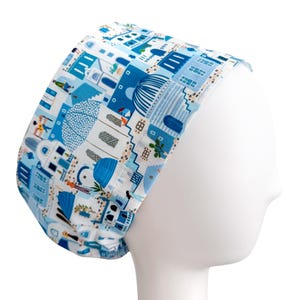 May include: A white and blue patterned scrub cap featuring a Mediterranean village design. The cap is made of fabric and is designed to fit comfortably. The pattern includes buildings, domes, and figures in shades of blue.