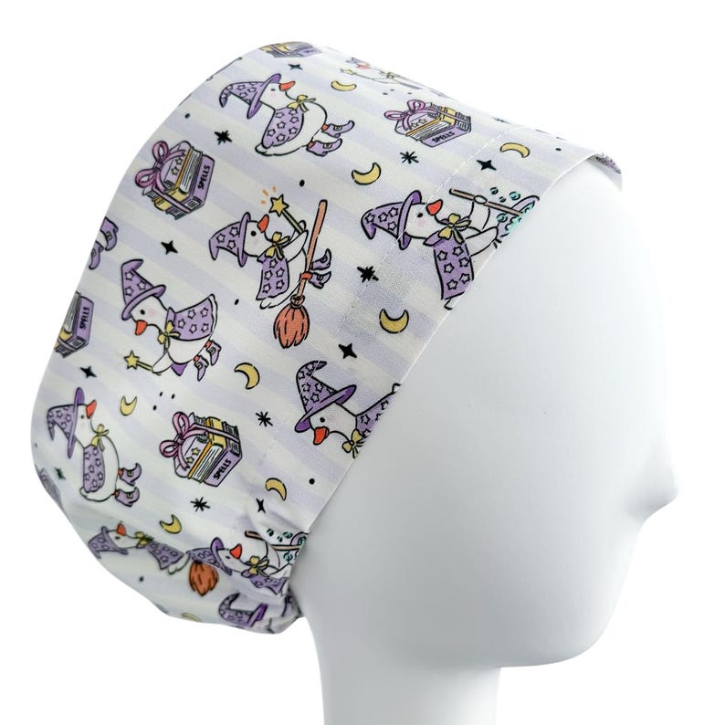 May include: A white and lavender striped scrub cap featuring a whimsical pattern of witch ducks. The ducks are depicted with purple witch hats, holding wands and riding brooms. Other elements include spell books, moons, and stars.