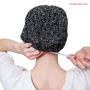 May include: A black and white patterned cap featuring a repeating cat face design. The cap is being adjusted with a white elastic band. The words "Example when worn" are visible in the upper right corner.