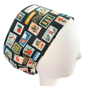 May include: A dark green scrub cap featuring a repeating pattern of colorful postage stamps. The stamps depict various images, including gifts, flowers, and skiers. The cap has the words "PAR AVION" and "FIRST CLASS POSTAGE" printed on it.