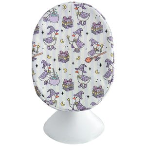 May include: A white, oval-shaped object with a whimsical pattern of cartoon ducks dressed as witches. The ducks wear purple hats and capes, holding wands and riding brooms. The background has vertical stripes, moons, stars, and gift boxes.