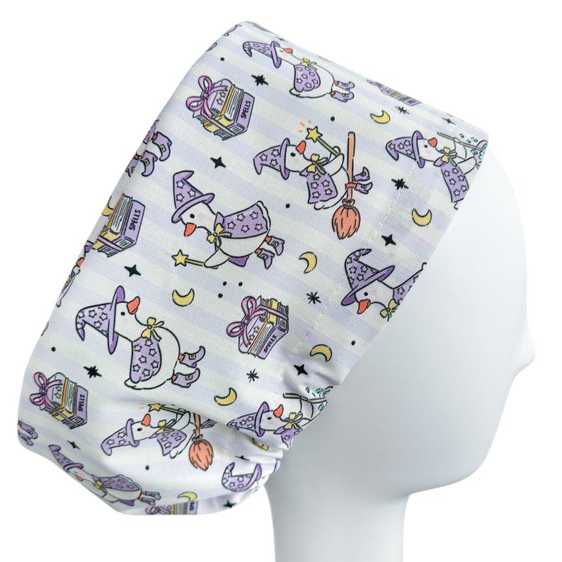 May include: A white scrub cap with a repeating pattern of cartoon geese dressed as witches, wearing purple hats and capes, flying on brooms, and holding wands. The background has vertical stripes and stars, moons, and spell books.