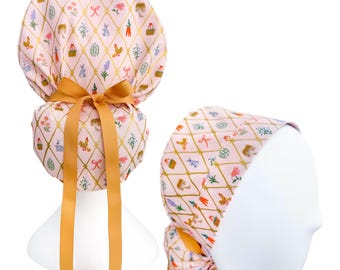Puede incluir: Un gorro quirúrgico rosa con un patrón repetido de ilustraciones con temática de Pascua, que incluye pollos, cestas y huevos, dentro de una cuadrícula de diamantes dorados. El gorro presenta un lazo dorado y cintas largas a juego.