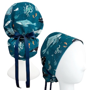 May include: A teal scrub cap featuring a pattern of whales, octopuses, and seahorses. The cap has a gathered back with a dark navy blue ribbon tie. The fabric displays various marine life illustrations in shades of white, light blue, and orange.