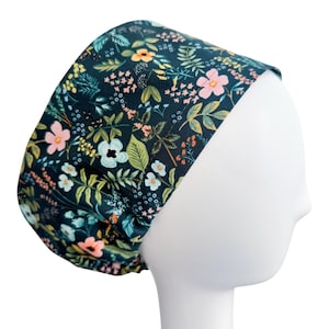 May include: A dark teal floral scrub cap featuring a pattern of pink, white, yellow, and orange flowers with green leaves. The cap is shown on a white mannequin head, showcasing the design and fit.
