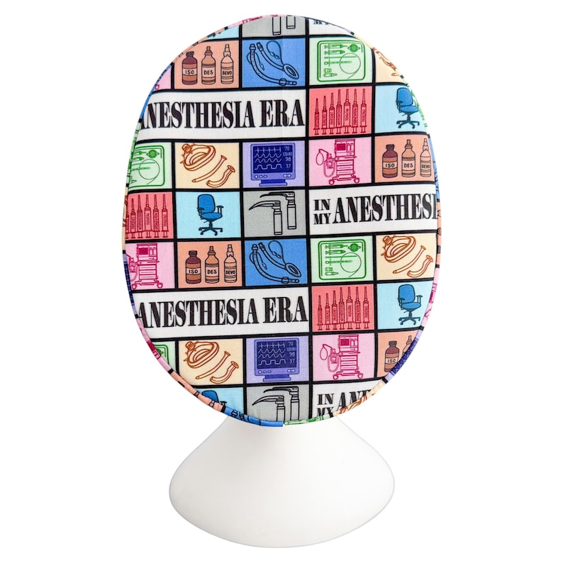 May include: A colorful surgical scrub cap with a repeating pattern of medical equipment and the text "ANESTHESIA ERA IN MY ANESTHESIA". The design includes images of syringes, monitors, and anesthesia supplies. The cap is displayed on a white stand.