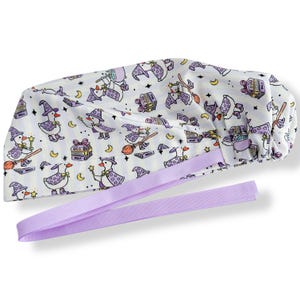 May include: White surgical scrub cap with a whimsical pattern of cartoon ducks dressed as witches, carrying wands and riding brooms. The cap has a lavender ribbon tie and is decorated with stars, moons, and spell books.