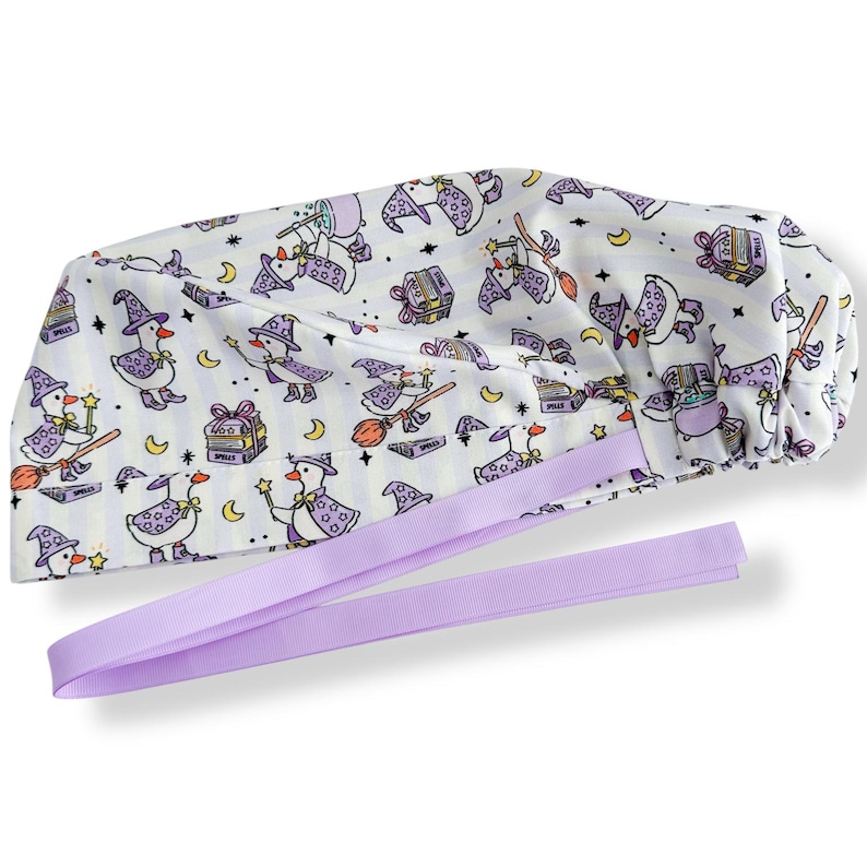 May include: White surgical scrub cap with a whimsical pattern of cartoon ducks dressed as witches, carrying wands and riding brooms. The cap has a lavender ribbon tie and is decorated with stars, moons, and spell books.