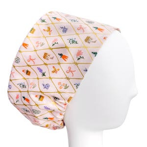 May include: A pink scrub cap with a gold diamond pattern and colorful floral and Easter-themed illustrations. The cap features images of flowers, baskets, chickens, and bows. The cap is displayed on a white mannequin head.