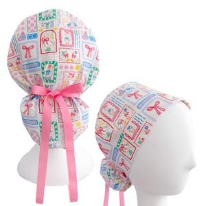 May include: A white scrub cap with a repeating pattern of framed ducks, bows, and hearts in pink, blue, and green. The cap has a pink ribbon tie. The cap is shown on a white mannequin head.