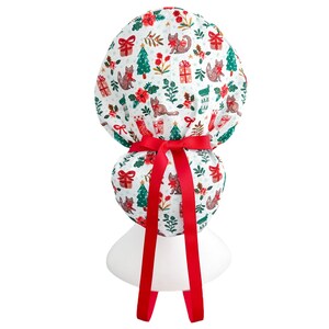 May include: A white scrub cap with a festive Christmas pattern featuring cats, gifts, Christmas trees, and floral accents. The cap is tied with a bright red ribbon. Designed for medical or professional use.