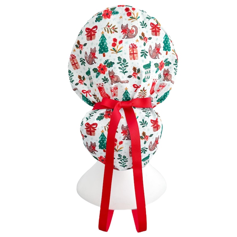 May include: A white scrub cap with a festive Christmas pattern featuring cats, gifts, Christmas trees, and floral accents. The cap is tied with a bright red ribbon. Designed for medical or professional use.