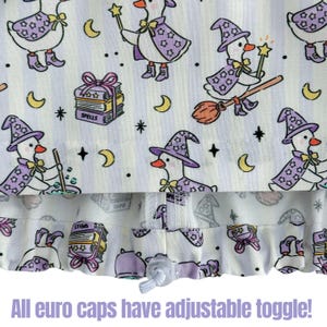 May include: White fabric with a repeating pattern of cartoon geese dressed as witches, wearing purple hats and capes. The design includes moons, stars, and spell books. The text "All euro caps have adjustable toggle!" is at the bottom.