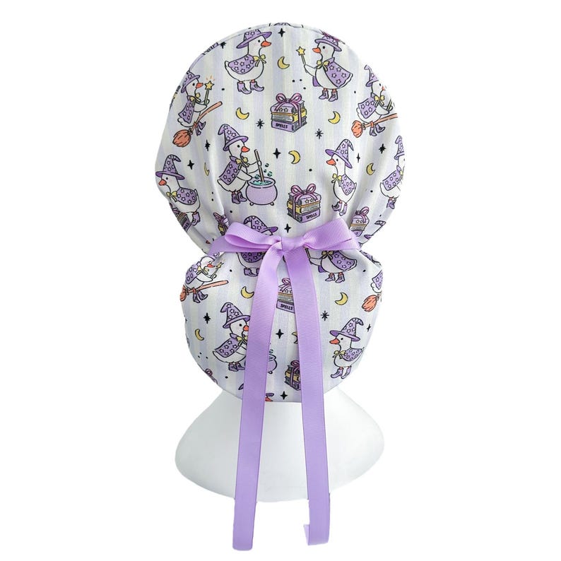 May include: A white scrub cap with a repeating pattern of witch ducks, moons, stars, and gift boxes. The cap is tied with a lavender bow and ribbons. The witch ducks wear purple hats and cloaks.