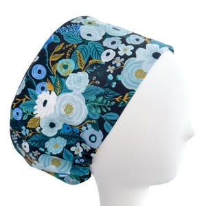 May include: A floral patterned scrub cap featuring a dark navy background with light blue, white, and gold flowers and leaves. The cap is designed to fit comfortably and securely, with a gathered back and a wide headband.