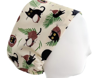 May include: A cream-colored scrub cap featuring a repeating pattern of black cats, coconuts, and green palm fronds. The cats have yellow eyes and are in various poses. The coconuts are brown. The cap is designed to fit over the head.