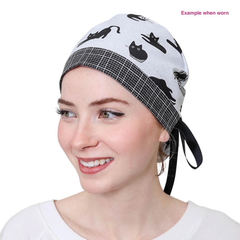 May include: A white surgical scrub cap featuring a black cat design. The cap has a black and white chequered border and black ties. The text "Example when worn" is visible in the upper right corner.