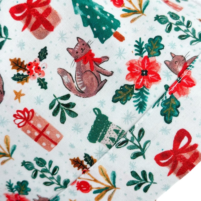 May include: White fabric featuring a festive pattern of Christmas illustrations. The design includes red gift boxes with bows, green Christmas trees, brown cats with red scarves, red poinsettias, and green sprigs.