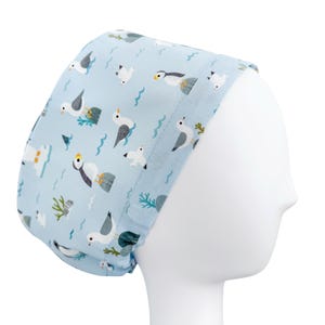 May include: A light blue surgical scrub cap featuring a repeating pattern of cartoon seagulls, puffins, and ocean elements. The cap is made of fabric and is designed to cover the head and hair.