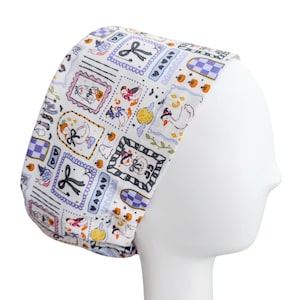 May include: A white scrub cap featuring a repeating pattern of framed illustrations including geese wearing hats, pumpkins, bows, and checkered squares in shades of purple, orange, and black. The fabric has a smooth texture and a fitted, rounded shape.