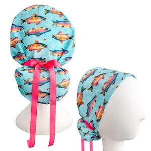 May include: A light blue scrub cap featuring a repeating pattern of colorful fish. The fish are in shades of orange, yellow, pink, and blue. The cap has a pink ribbon tie. A second cap is shown on a mannequin head.