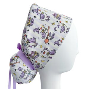 May include: A white and lavender scrub cap featuring a whimsical pattern of witch ducks with spell books, brooms, and stars. The cap has a striped background and a lavender ribbon tie. The cap is designed to be worn on the head.