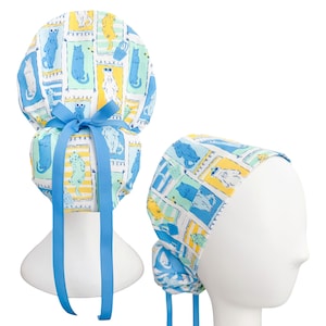 May include: A patterned scrub cap featuring a repeating design of cats in blue, yellow, and white on a light blue background. The cap has a blue ribbon tie and is displayed on a white mannequin head. The overall theme is cat-themed accessories.