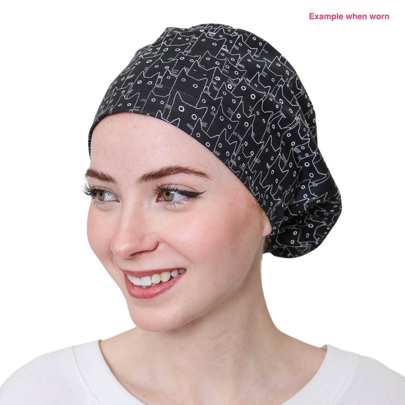 May include: Black surgical scrub cap with a repeating white cat face pattern. The cap is shown on a person. The text "Example when worn" is in the upper right corner.