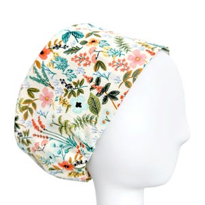 May include: A scrub cap featuring a cream-colored background with a vibrant botanical print. The pattern includes various flowers in shades of pink, orange, and light blue, interspersed with green leaves, ferns, and small dark berries across the fabric.