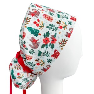 May include: A white scrub cap with a festive Christmas pattern featuring brown cats, red bows, green Christmas trees, and gift boxes. The cap has a red bow and ties at the back. The design is printed on a white background.