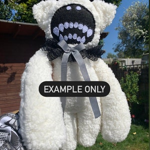 May include: White plush teddy bear with a black felt face and large white teeth. The bear is wearing a black and white lace collar with a gray satin bow. The text "EXAMPLE ONLY" is printed on the bear's chest.