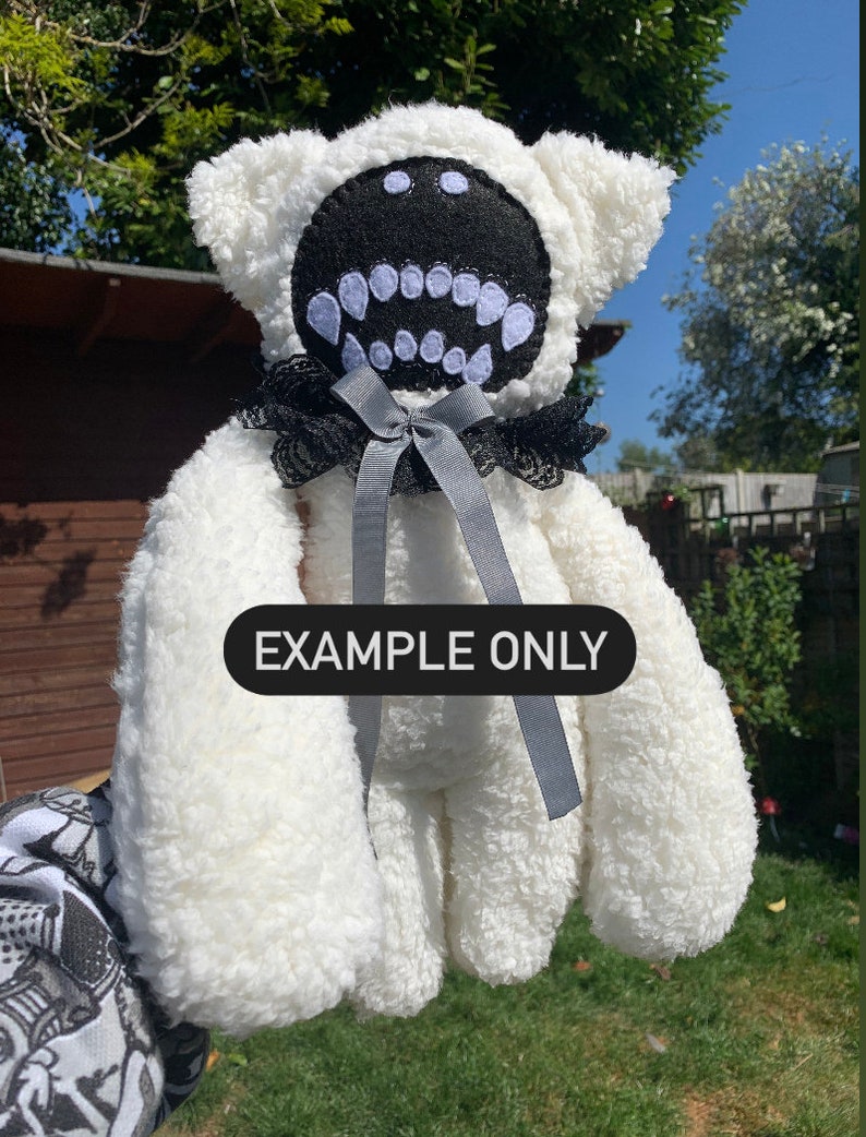 May include: White plush teddy bear with a black felt face and large white teeth. The bear is wearing a black and white lace collar with a gray satin bow. The text "EXAMPLE ONLY" is printed on the bear's chest.