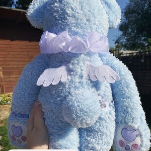 May include: A light blue plush teddy bear with white wings and a white satin bow around its neck. The bear has embroidered paw prints on its feet.