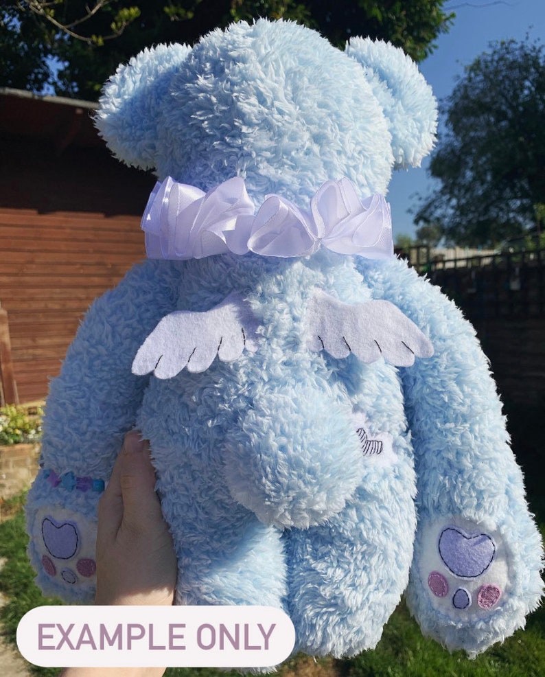 May include: A light blue plush teddy bear with white wings and a white satin bow around its neck. The bear has embroidered paw prints on its feet.