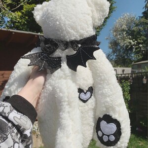 May include: White plush teddy bear with black bat wings, a black bow, and a white heart and paw print patches.  The bear is being held by a hand.