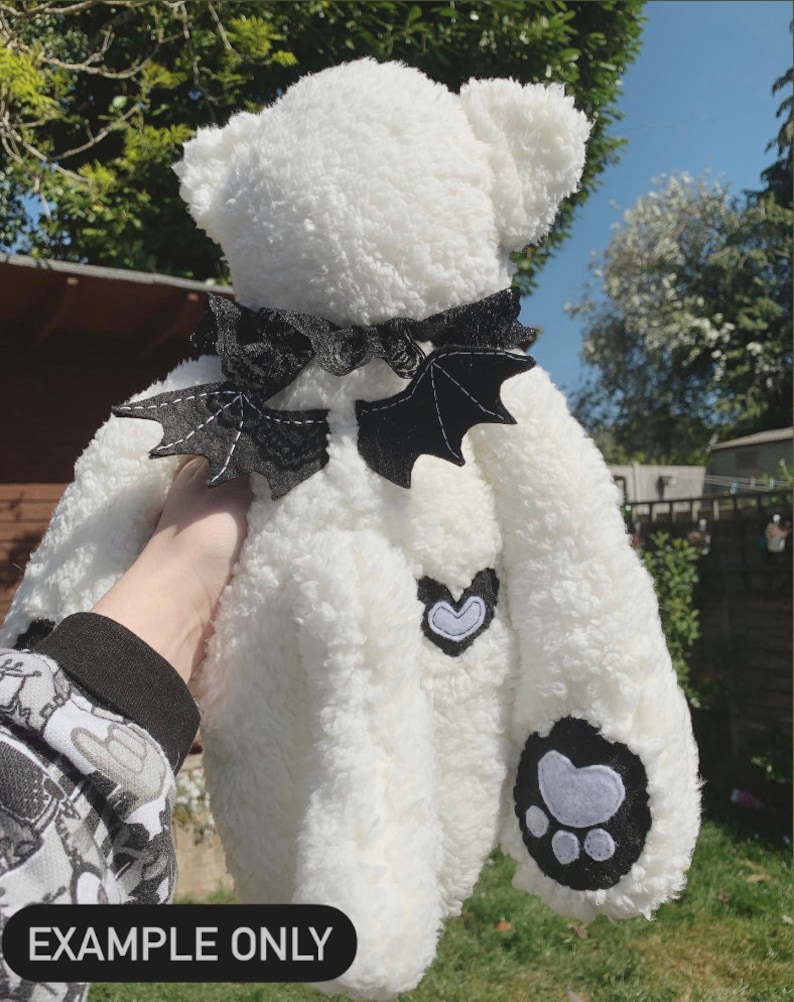 May include: White plush teddy bear with black bat wings, a black bow, and a white heart and paw print patches.  The bear is being held by a hand.