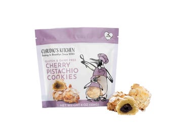 May include: A purple bag of gluten and dairy free cherry pistachio cookies. The bag has a cartoon penguin on it. The bag says "Claudio's Kitchen Baking in Brooklyn Since 2020". There are two cookies in front of the bag, one is broken open to show the cherry filling.