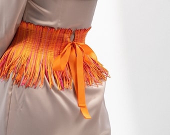 Pode incluir: Um vestido de cetim bege com um cinto de franjas coloridas amarrado com uma fita laranja brilhante.